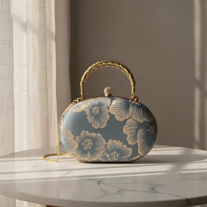 Peut inclure: Une pochette de forme ovale dans une teinte bleu sourd, ornée d'un motif floral doré. Le sac possède une poignée en métal doré texturé et un petit fermoir rond incrusté de cristaux sur le dessus. Une fine chaîne dorée est fixée sur le côté.