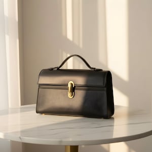 K&ouml;nnte beinhalten: Eine schwarze Lederhandtasche mit einem oberen Griff und einem goldfarbenen Verschluss. Die Tasche hat ein strukturiertes Design und eine strukturierte Oberfl&auml;che. Die Handtasche steht auf einem wei&szlig;en Marmortisch, mit Schatten durch Sonnenlicht.