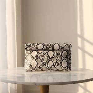 Può includere: Pochette rettangolare con motivo a pelle di serpente nei toni del nero, bianco e grigio. La borsa è ripiegata nella parte superiore e poggia su un tavolo di marmo bianco.