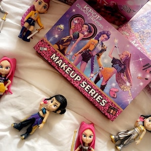May include: A selection of KPOP Demon Hunters dolls and makeup series packaging. The dolls are in various poses and outfits, with pink and purple accents. The makeup series box is pink with the words "MAKEUP SERIES".
