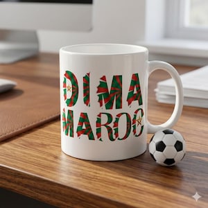 May include: A white ceramic mug featuring the text DIMA MAROC in a bold, stylized font with a red and green sunburst pattern. The letter D and the letter O each contain a small yellow five-pointed star. A miniature black and white soccer ball.
