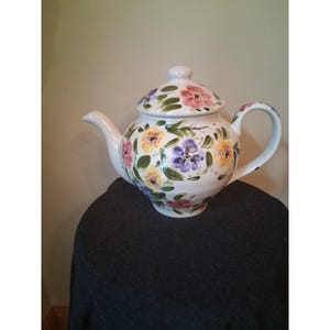 Peut inclure: Une théière en céramique blanche ornée d'un motif floral peint à la main avec des fleurs roses, jaunes et violettes accentuées par des feuilles vertes. La théière comprend un couvercle assorti avec un bouton, un bec verseur et une anse.