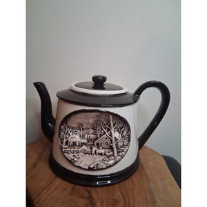 May include: A ceramic teapot featuring a cream-coloured body with a dark brown lid, handle, spout, and base. An oval relief panel on the front depicts a detailed winter landscape with a house, bare trees, and figures in a snowy outdoor setting.
