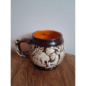 May include: A brown and cream ceramic mug with a handle. The mug has a relief design of a man, grapes, and vines. The interior of the mug is orange.