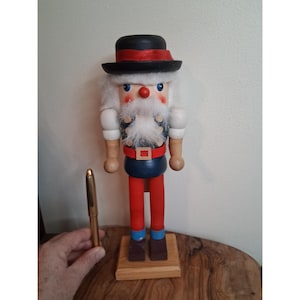 May include: A wooden nutcracker figurine with a black hat, red trim, and a white beard. The figurine has a red nose, blue eyes, and is wearing a blue vest, red trousers, and brown shoes. The nutcracker is approximately 30 cm tall.