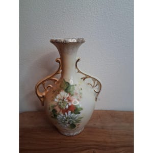 May include: Cream ceramic vase with gold detailing and a floral pattern. The vase has two decorative handles and a flared rim. The flowers are painted in pink, orange, and white with green leaves. A decorative item for the home.