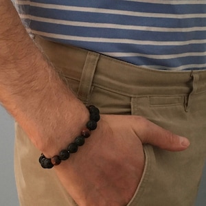 Könnte beinhalten: Ein braunes Armband mit schwarzen und roten Perlen. Das Armband wird am Handgelenk einer Person getragen, die ein blau-weiß gestreiftes Hemd und eine Khakihose trägt.