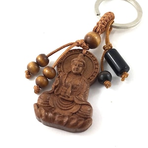 Puede incluir: Un llavero de Buda de madera con un llavero plateado. La figura de Buda marrón está tallada con detalle. El llavero también presenta cuentas de madera y una cuenta cilíndrica negra, todo conectado con un cordón marrón.