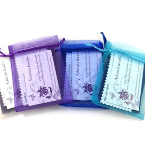 May include: Three organza bags in purple, blue and turquoise, each containing a white card. The cards feature a butterfly design and the text "Ath&eacute;na&iuml;s Jewelry" with the website address and a description of the jewellery.