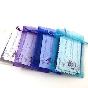 Pu&ograve; includere: Quattro sacchetti di organza in tonalit&agrave; di viola, blu e turchese, ognuno con un cartoncino bianco all'interno. I cartoncini presentano un disegno di farfalla e il testo "Ath&eacute;na&iuml;s Jewelry" e un indirizzo di sito web.