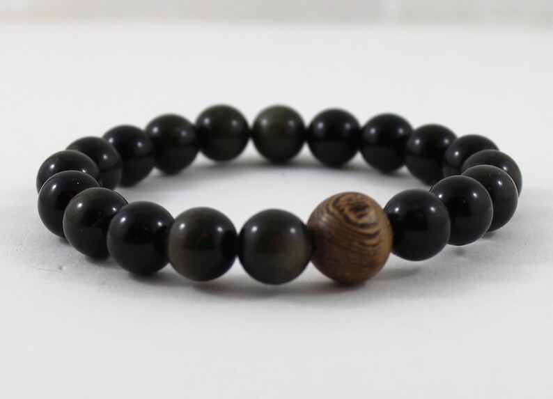 May include: A black beaded bracelet with a single brown wooden bead. The bracelet is made of round beads and is approximately 18 cm in length.