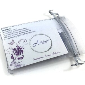 May include: A white card with purple floral designs and text that reads "Obsidian: A powerful stone A strongly protective stone A powerful stone in all senses Relieves tension, brings clarity Promotes strength and compassion Ath&eacute;nais Jewelry Inspiration. Energy Balance." The card is in a white and grey organza bag.