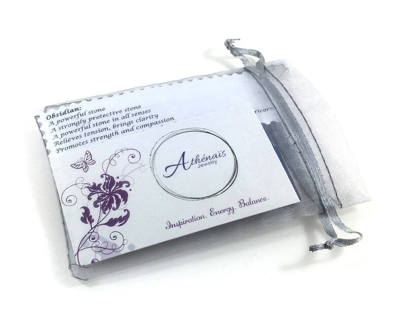May include: A white card with purple floral designs and text that reads "Obsidian: A powerful stone A strongly protective stone A powerful stone in all senses Relieves tension, brings clarity Promotes strength and compassion Ath&eacute;nais Jewelry Inspiration. Energy Balance." The card is in a white and grey organza bag.
