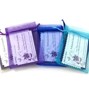 Pu&ograve; includere: Tre sacchetti di organza viola, blu e turchese, ognuno contenente una carta bianca. Le carte presentano un design a farfalla e il testo "Ath&eacute;na&iuml;s Jewelry" con l'indirizzo del sito web e una descrizione dei gioielli.