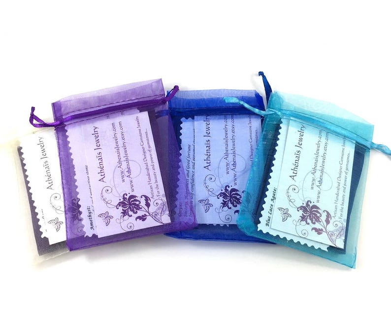 Pu&ograve; includere: Tre sacchetti di organza viola, blu e turchese, ognuno contenente una carta bianca. Le carte presentano un design a farfalla e il testo "Ath&eacute;na&iuml;s Jewelry" con l'indirizzo del sito web e una descrizione dei gioielli.