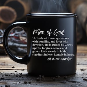 May include: Black ceramic mug featuring the text "Man of God" and a description of a man's qualities. The mug also includes the phrase "He is my Grandpa". The mug is on a wooden surface.