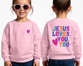 Peut inclure: Un sweat-shirt rose clair avec un petit cœur rose sur le devant et l'inscription "JESUS LOVES YOU, TOO" en lettres colorées au dos. Le sweat-shirt est porté par un enfant portant des lunettes de soleil.