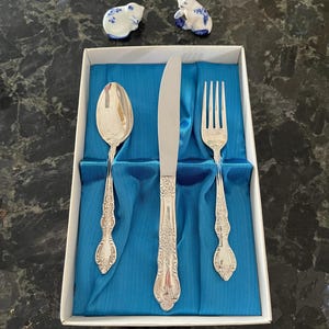 Könnte beinhalten: Ein silberfarbenes Besteckset, bestehend aus einem Löffel, Messer und Gabel, präsentiert in einer weißen Box mit blauem Satinfutter. Zwei kleine Porzellanfiguren von Tieren mit blauen Blumenmustern sind ebenfalls zu sehen.