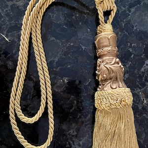 May include: A gold-toned curtain tieback with a decorative tassel. The tieback has a twisted rope design leading to an ornate, carved finial and a long, flowing tassel. The design is elegant and suitable for home decor.