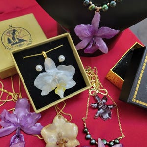 May include: A collection of jewelry featuring orchid-shaped pendants in purple, white, and dark tones, paired with gold-tone chains and pearl accents. The items are displayed with branded boxes featuring a compass logo and the text GAIN COMPASS.