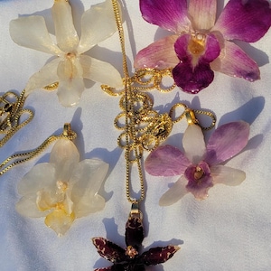 May include: Five orchid flower pendants attached to gold-toned metal chains. Two pendants are white, one is deep purple with white speckles, one is light purple, and one is a mix of white and purple. Each flower has a gold-toned metal bail at the top.