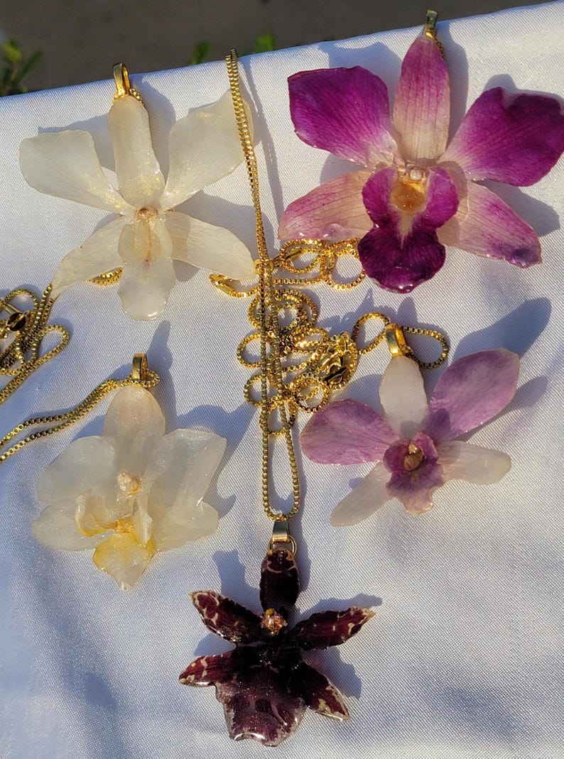 May include: Five orchid flower pendants attached to gold-toned metal chains. Two pendants are white, one is deep purple with white speckles, one is light purple, and one is a mix of white and purple. Each flower has a gold-toned metal bail at the top.