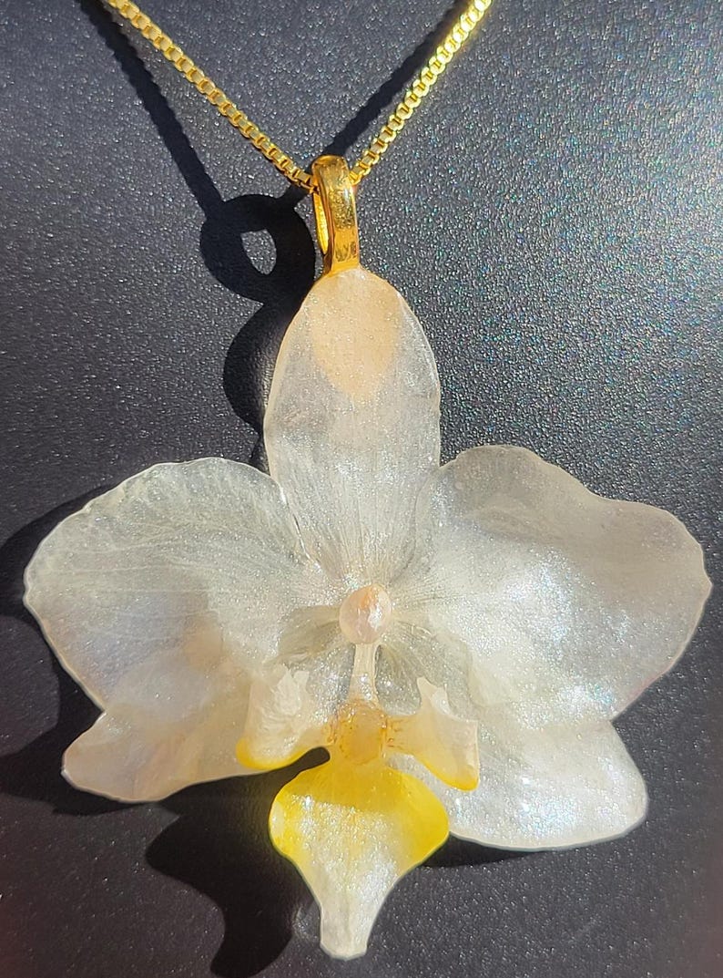 May include: A pendant shaped like an orchid flower with translucent white petals featuring a subtle pearlescent shimmer. The center of the flower displays a bright yellow lip and a small pale pink column. The pendant hangs from a gold-toned chain.
