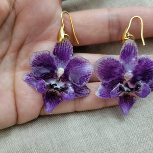 May include: A pair of dangle earrings featuring realistic purple orchid flower charms with white edges and dark purple centers. The orchid petals have a glossy finish and are attached to gold-toned metal ear wires with a simple hook design.