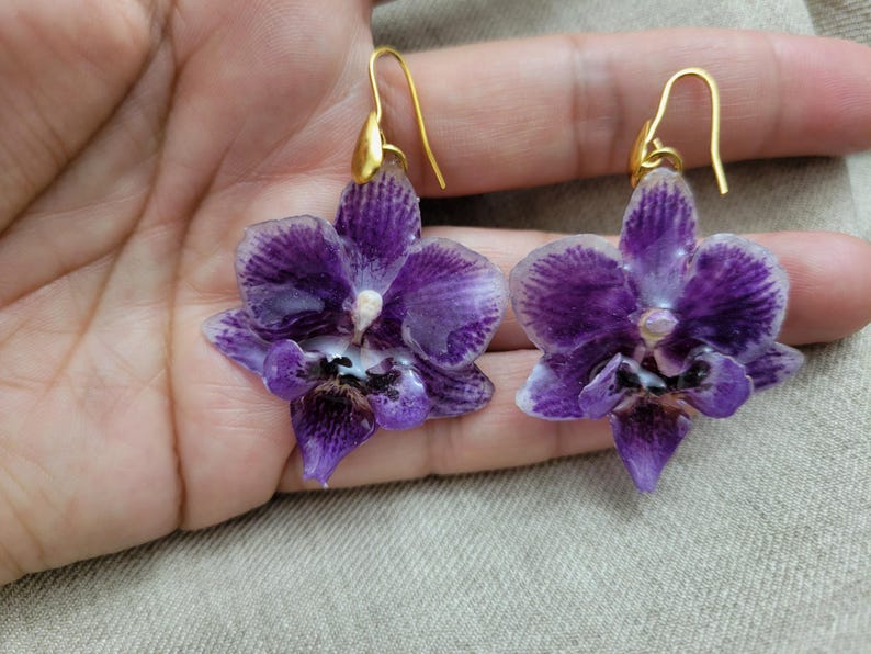 May include: A pair of dangle earrings featuring realistic purple orchid flower charms with white edges and dark purple centers. The orchid petals have a glossy finish and are attached to gold-toned metal ear wires with a simple hook design.