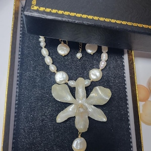 May include: A necklace and earring set featuring white freshwater pearls of varying shapes and sizes. The necklace includes a large, translucent, off-white orchid-shaped pendant with a single pearl drop hanging from the center of the flower.
