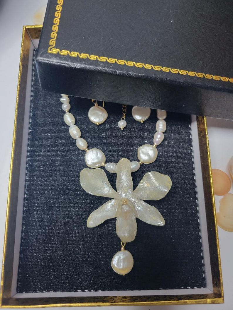 May include: A necklace and earring set featuring white freshwater pearls of varying shapes and sizes. The necklace includes a large, translucent, off-white orchid-shaped pendant with a single pearl drop hanging from the center of the flower.