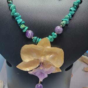May include: A necklace and earring set featuring turquoise chips and round purple amethyst beads. The necklace includes a large, central gold and purple orchid-shaped pendant. The earrings consist of a single purple bead and a turquoise bead.