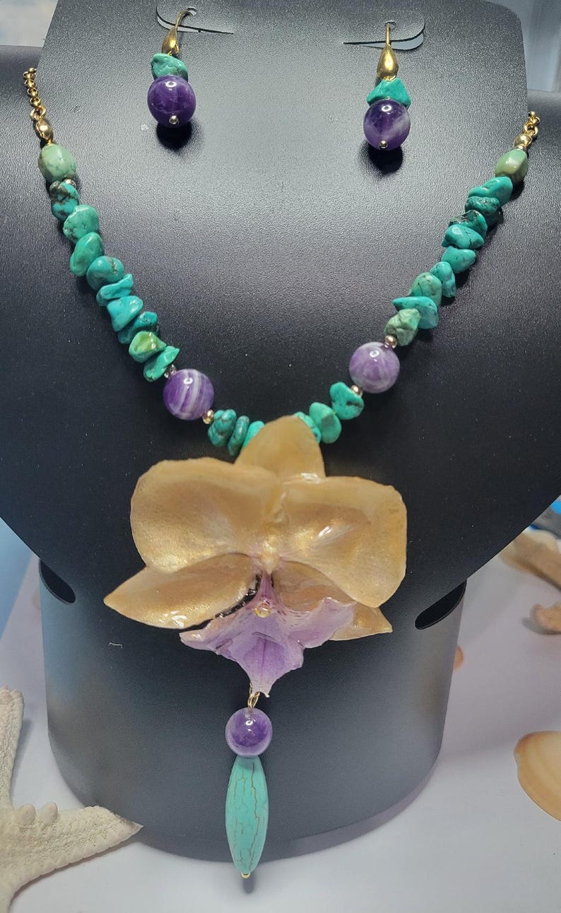May include: A necklace and earring set featuring turquoise chips and round purple amethyst beads. The necklace includes a large, central gold and purple orchid-shaped pendant. The earrings consist of a single purple bead and a turquoise bead.