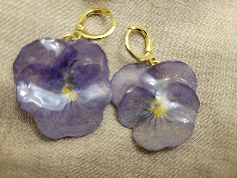 May include: A pair of earrings featuring pressed purple pansy flowers encased in a clear, glossy resin. Each flower has a yellow center and is attached to a gold-toned metal hoop clasp. The petals show natural veining and translucent purple hues.