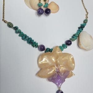 May include: A jewelry set featuring a necklace and matching earrings. The necklace has a gold-tone chain with turquoise chips, purple amethyst beads, and a large, glossy, translucent peach and purple orchid-shaped pendant with a turquoise drop.