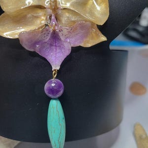 May include: A pendant featuring a translucent gold and purple orchid flower attached to a gold-tone jump ring. Below the flower hangs a round, smooth purple bead followed by a long, oval-shaped turquoise bead with brown veining throughout.