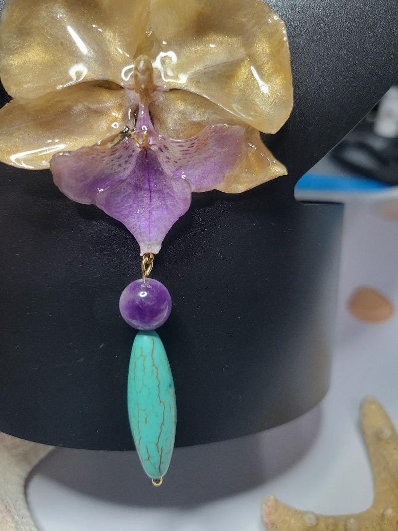 May include: A pendant featuring a translucent gold and purple orchid flower attached to a gold-tone jump ring. Below the flower hangs a round, smooth purple bead followed by a long, oval-shaped turquoise bead with brown veining throughout.