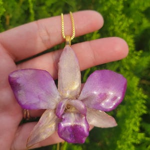 May include: A pendant shaped like an orchid flower featuring a glossy finish with blended shades of purple, cream, and gold metallic accents. The orchid is attached to a thin gold-toned metal chain that connects to a small gold-colored bail.
