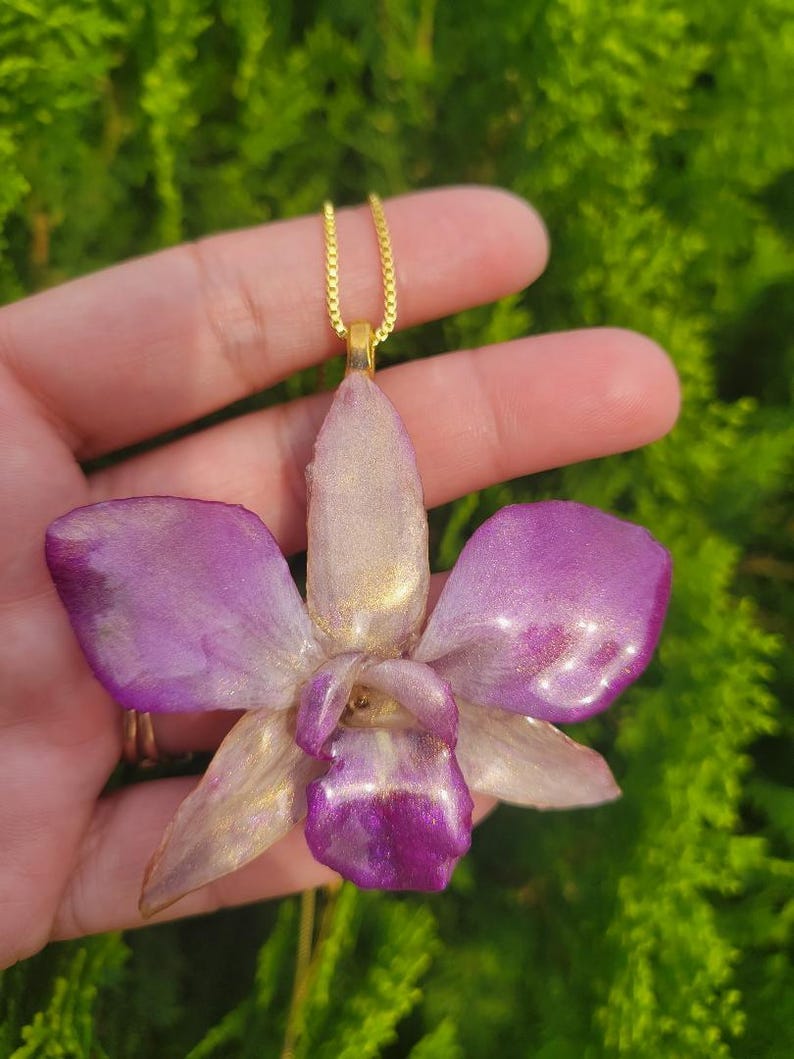 May include: A pendant shaped like an orchid flower featuring a glossy finish with blended shades of purple, cream, and gold metallic accents. The orchid is attached to a thin gold-toned metal chain that connects to a small gold-colored bail.