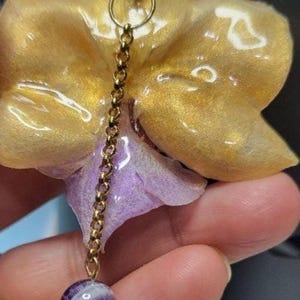 May include: A pendant necklace featuring a gold-colored orchid flower with a purple center, suspended from a gold-tone chain. The design includes a round purple and white striped bead, turquoise-colored stone beads, and a gold-colored metal jump ring.