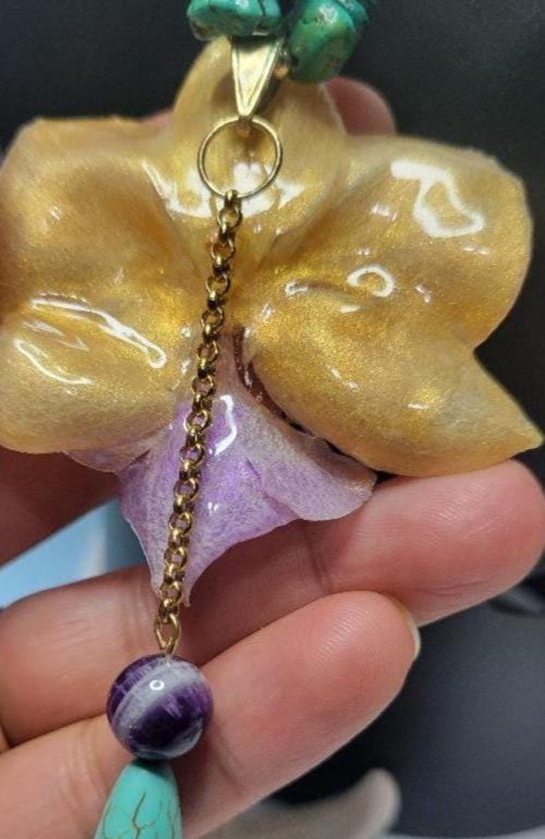 May include: A pendant necklace featuring a gold-colored orchid flower with a purple center, suspended from a gold-tone chain. The design includes a round purple and white striped bead, turquoise-colored stone beads, and a gold-colored metal jump ring.