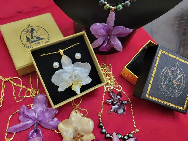 May include: A collection of jewelry featuring orchid-shaped pendants in purple, white, and dark tones, paired with gold-tone chains and pearl accents. The items are displayed with branded boxes featuring a compass logo and the text GAIN COMPASS.