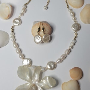 May include: A necklace and matching pair of drop earrings featuring white freshwater pearls of varying shapes and sizes. The necklace includes a large, translucent, flower-shaped pendant and a gold-toned chain with a lobster claw clasp.