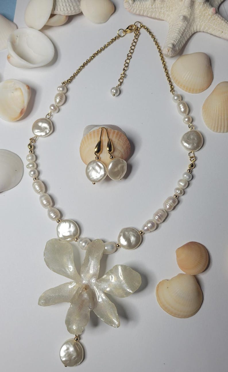May include: A necklace and matching pair of drop earrings featuring white freshwater pearls of varying shapes and sizes. The necklace includes a large, translucent, flower-shaped pendant and a gold-toned chain with a lobster claw clasp.