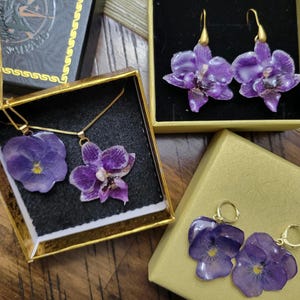 May include: A jewelry set featuring purple flower pendants and earrings. One necklace has a pansy and an orchid pendant on a gold chain. One pair of orchid drop earrings and one pair of pansy hoop earrings are displayed in gold-colored gift boxes.