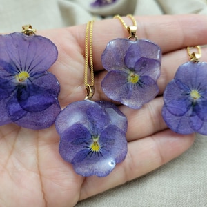 May include: Four pendants featuring preserved purple pansy flowers encased in a clear, glossy resin. Each flower has a yellow center and is attached to a thin gold-toned metal chain. The petals display natural veining and deep violet hues.