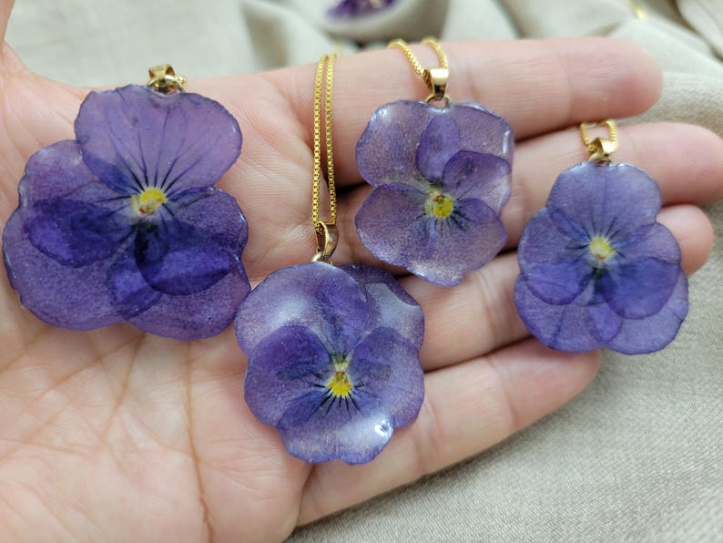 May include: Four pendants featuring preserved purple pansy flowers encased in a clear, glossy resin. Each flower has a yellow center and is attached to a thin gold-toned metal chain. The petals display natural veining and deep violet hues.