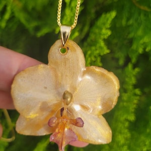 May include: A gold-toned orchid flower pendant necklace featuring a glossy, resin-coated finish with shimmering gold accents and a small, deep purple center detail, suspended from a delicate gold-colored metal chain.