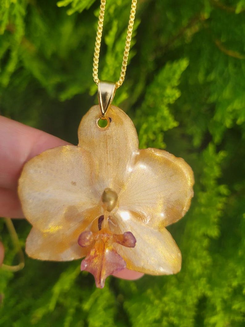 May include: A gold-toned orchid flower pendant necklace featuring a glossy, resin-coated finish with shimmering gold accents and a small, deep purple center detail, suspended from a delicate gold-colored metal chain.