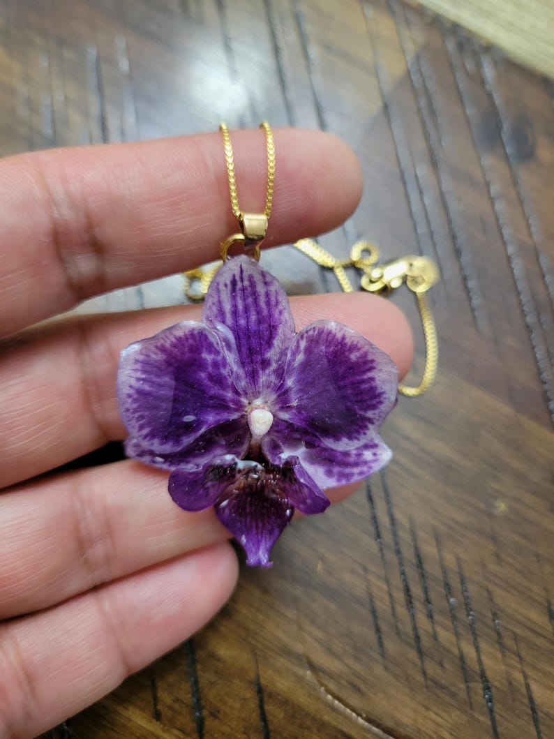 May include: A pendant necklace featuring a preserved purple orchid flower with white edges and a dark center, suspended from a thin gold-tone chain. The orchid petals have a glossy, resin-like finish, showcasing natural veining and color gradients.